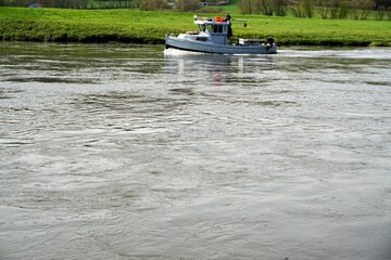 Kleines weißes Schiff auf fährt über Fluss vor grüner Wiese bei Sonne am Nachmittag im Frühling