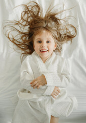 Joyful girl in a white bathrobe lies in bed, looks at the camera. - obrazy, fototapety, plakaty
