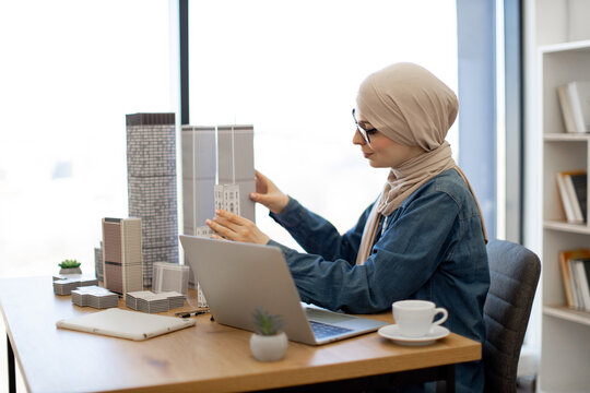Thoughtful muslim lady in hijab placing architectural models on desk surface near smart gadgets in creative workplace. Residential architect analysing project data using wireless technologies.
