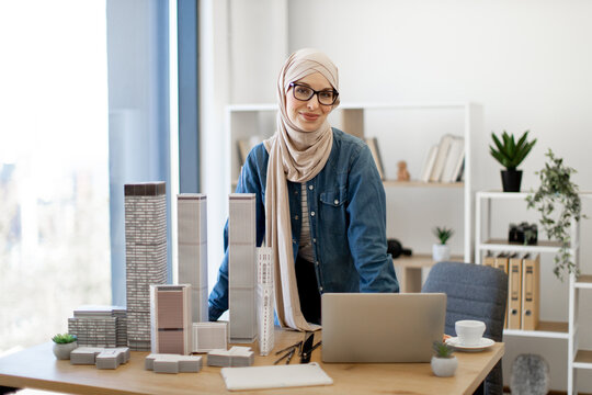 Adult arabian female in hijab posing behind writing desk with gadgets and maquettes in architecture office. Experienced professional having coffee break from project management in creative workplace.