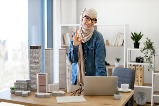 Adult arabian female in hijab posing behind writing desk with gadgets and maquettes in architecture office. Experienced professional having coffee break from project management in creative workplace.