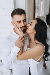 Stylish bearded groom and handsome brunette bride hugging, kissing while standing near the window of a white brick wall in the interior, indoors. Wedding photography, close-up portrait.