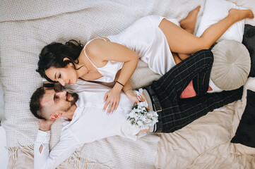 Stylish bearded groom and beautiful brunette bride with bouquet hugging while lying on bed in interior, indoors. Wedding photography, portrait.