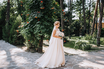 A beautiful young blonde model bride in a white lace dress with a crown, a diadem stands in a park, forest in nature with a bouquet in her hands. Wedding portrait, photography.