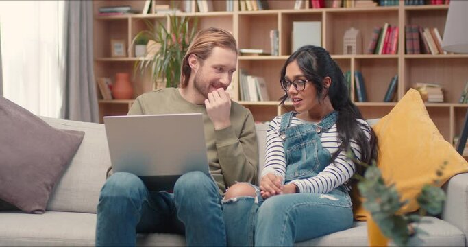 Beautiful multiethnic friends spending free time together in library. Handsome man holding laptop computer and sitting on a comfortable sofa with young woman. Positive people talking or gossiping.