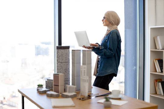 Arabian businesswoman wearing hijab and glasses standing with laptop in hands near panoramic window in creative workplace. Young architect carrying out city complex construction plan using gadgets.