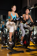 Fototapeta na wymiar Two young adult women taking indoor cycling class at fitness center, doing cardio riding bike