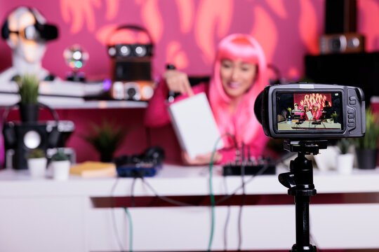 Asian vlogger filming white box review in recording studio talking with subscribers using professional vlogging camera. Content creator with pink hair talking with subscribers during live streaming