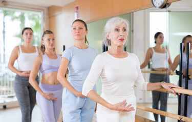 Slim aged woman and other attendees practicing ballet at ballet barre in dance studio during training session