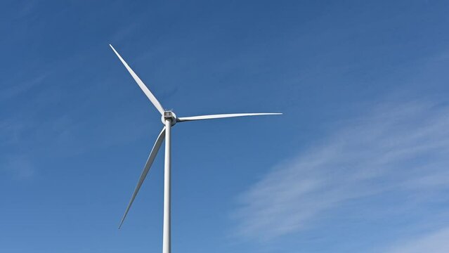 A large wind turbine rotates but gradually slows to a stop. Blue sky with wispy clouds behind.