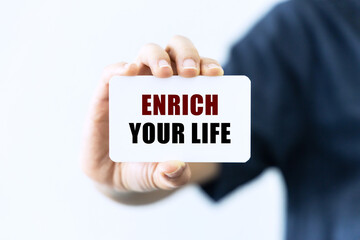 Enrich your life text on blank business card being held by a woman's hand with blurred background. Business concept about enriching your life.