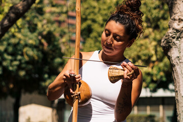 mujer morena tocando berimbau instrumento de percusión de capoeira, en un parque con un día...