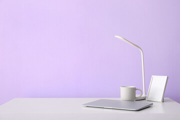 Workplace with laptop, cup, blank frame and lamp near lilac wall
