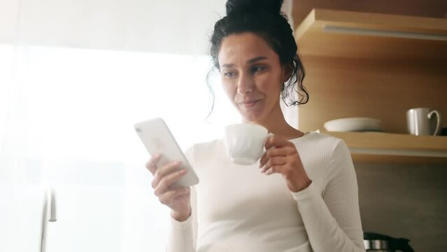 Pretty hispanic woman enjoying beautiful morning with cup of coffee using smartphone scrolling social media at sunny kitchen Charming young female relaxing and chatting on mobile phone at home