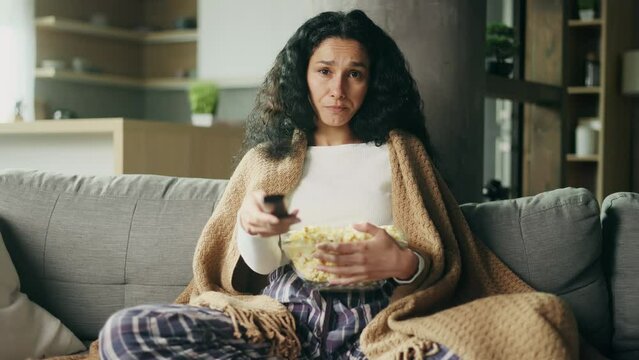 Camera view of tired young hispanic woman with remote controller switches channels on TV trying to choose something interesting at home Boring TV shows films movie 