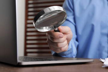 Man holding magnifier near laptop at wooden table, closeup. Online searching concept