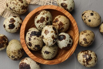 Wooden bowl and many speckled quail eggs on grey table, flat lay