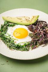 Plate with tasty fried egg, avocado and greens on green background