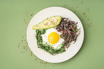 Plate with tasty fried egg, avocado and greens on green background
