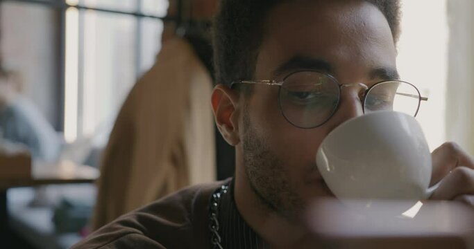 Close-up portrait of joyful African American student drinking coffee and using smartphone in cafe in daytime. Beverage and young people concept.