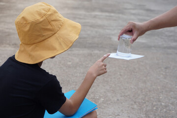 Student boy wears hat, hold book, learning science experiment about air pressure from glass of water which covered by paper. Concept, science subject activity, education. Learning by doing. Observe.  