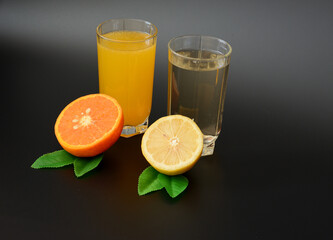 Two glasses with different citrus juices on a black background, next to them are halves of a lemon and an orange with leaves.