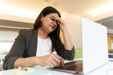 Stressed Asian mature woman holding head in hands, feeling headache after hardworking on laptop