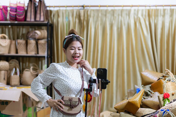 A beautiful Southeast Asian woman wearing a white dress is carrying a handicraft woven bag while doing a live broadcast of selling products through a social media app on her mobile phone.