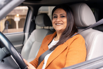 Confident Asian mature woman sitting at driver's seat with fasten belt, smiling looking at camera