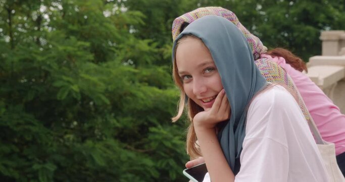 Religious smiling blonde cute muslim girl in grey headscarf looking at camera. Young teen girl walks in summer park with mom admires nature views. Customs, observance of Islamic religious traditions.