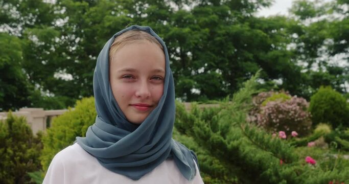 Portrait of smiling blonde muslim girl in grey headscarf looking at camera on nature. Laughing teen girl walks in summer park. Happy young shy modest female. Customs, observance of Islamic traditions.