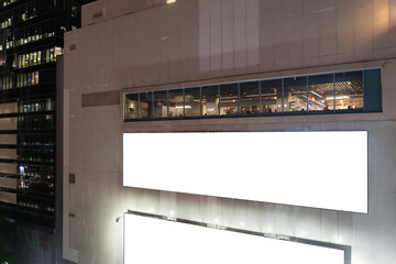 Clean white billboard on shiny glass building exterior with reflections at night time. Mock up. Large billboards for advertising buildings at the downtown economy.