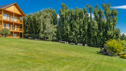 Fototapeta na wymiar Green trees and shrubs grow on the lawn on the hillside. Wooden cottage with balconies against the blue sky. Argentina. 
