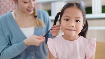 Happy loving family Asian mother is braiding hair her daughter's sitting on sofa at living room....