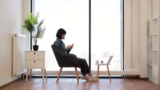 Pleasant dark haired woman typing messages on smartphone while sitting in comfy office chair. Caucasian female employee in eyeglasses chatting with coworkers online using modern gadget.