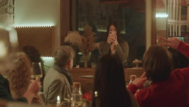 Asian woman takes photo and selfie with friends in modern gastro cafe using phone. Group of diverse people spend weekend evening, hangout on birthday party in restaurant. Concept of public catering.
