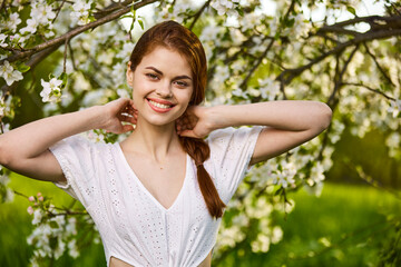 Fototapeta premium portrait of a joyful woman in a light dress against the background of a flowering tree, raising her hands to her head