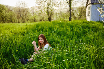 cheerful woman sitting in tall grass near small house in countryside