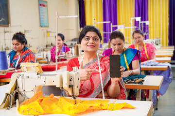 Indian women showing smartphone screen while working on sewing machine at factory