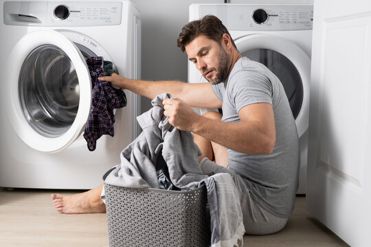 Man with clothes near washing machine. Handsome man sits in front of washing machine. Loads washer with dirty laundry. Man cleaning clothes. Housework for single alone guy. Home laundry.