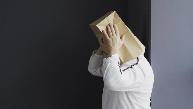 A sad man in a white shirt with a bag on his head, with a sad smiley face drawn, holds his head. Emotions and gestures.