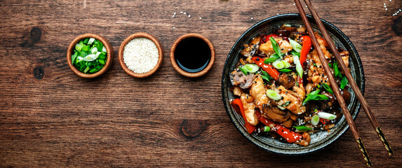 Stir fry chicken with paprika, mushrooms, green chives and sesame seeds in ceramic bowl.  Asian cuisine dish. Wooden kitchen table background, top view banner - obrazy, fototapety, plakaty