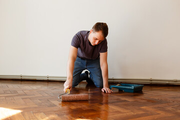 man varnishes oak parquet with a roller during renovation