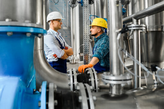 Two young adult workers of the plant talk amicably among engineered metal structures and pipelines.