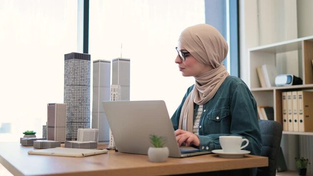 Focused bespectacled muslim female in hijab typing on portable computer at office desk with building models on it. Experienced architect developing urban design using modern gadgets and drawing tools.