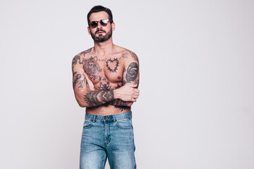 Fototapeta premium Portrait of handsome confident stylish hipster lambersexual model in jeans. Sexy modern man. Naked torso with tattoos. Fashion male posing in studio. Isolated on white. In sunglasses