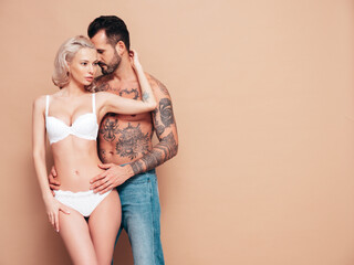 Sexy beautiful woman and her handsome boyfriend. Young passionate couple hugging. Man with nude tattoo torso. Blond model in lace lingerie wearing pure underwear. Posing in studio, near beige wall