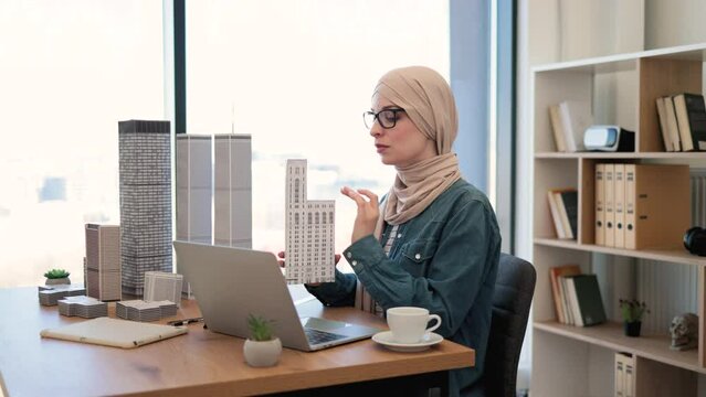 Attractive arabian businesswoman wearing hijab and holding multi-storey building model while seating at writing desk with digital devices on it. Modern developer reviewing plan for urban space.