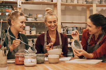 A company of three cheerful young women friends are painting ceramics in a pottery workshop. - obrazy, fototapety, plakaty