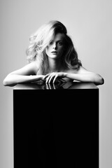Beauty, fashion and make-up concept. Beautiful sexy blonde woman with big wavy hair holding hands on black glass cube with copy space. Minimalistic and futuristic looking style. Black and white image
